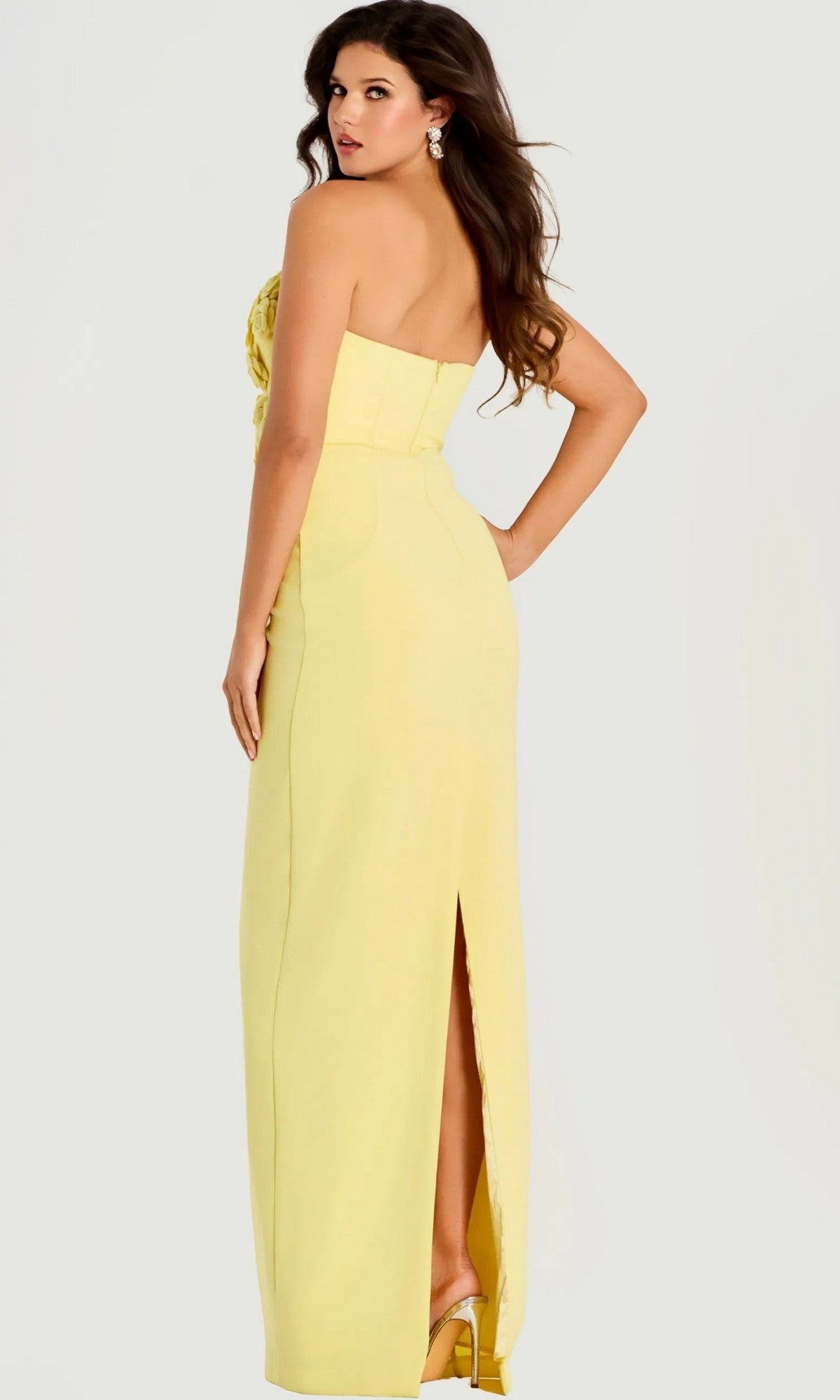 A woman poses against a plain background, looking over her shoulder in the Jovani 46884 long prom dress—a strapless yellow column gown featuring a back slit.